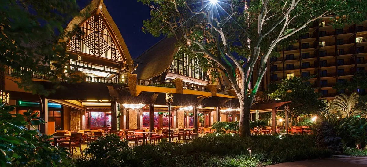A beautifully lit restaurant with a thatched roof and intricate wooden architecture is surrounded by lush greenery at night. Numerous tables and chairs are arranged under soft, warm lighting, creating an inviting outdoor dining area.
