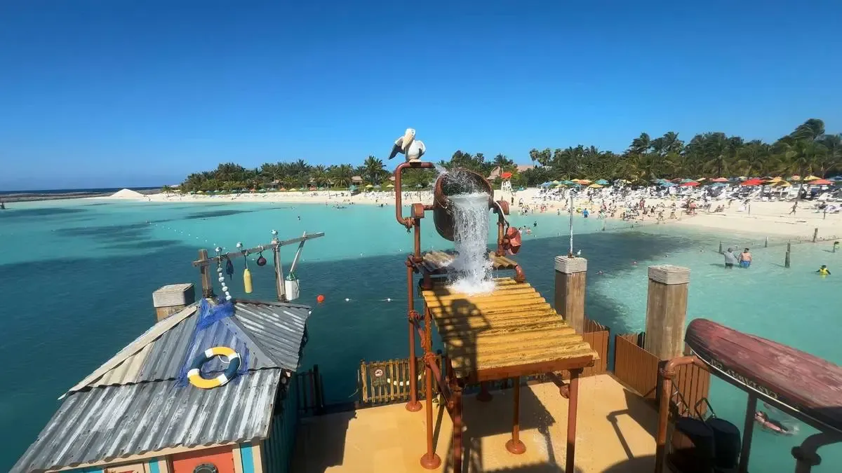 A vibrant beach scene shows a playful water feature with a large bucket pouring water, set against a backdrop of turquoise water and a sandy beach filled with colorful umbrellas and sunbathers. The sky is clear and blue, and palm trees line the beach, creating a tropical atmosphere.