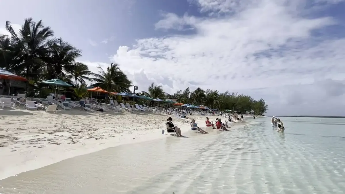 A serene beach scene with people relaxing on lounge chairs along the sandy shore, shaded by colorful umbrellas. Clear, shallow water gently laps against the beach, while palm trees sway in the background under a partly cloudy sky.
