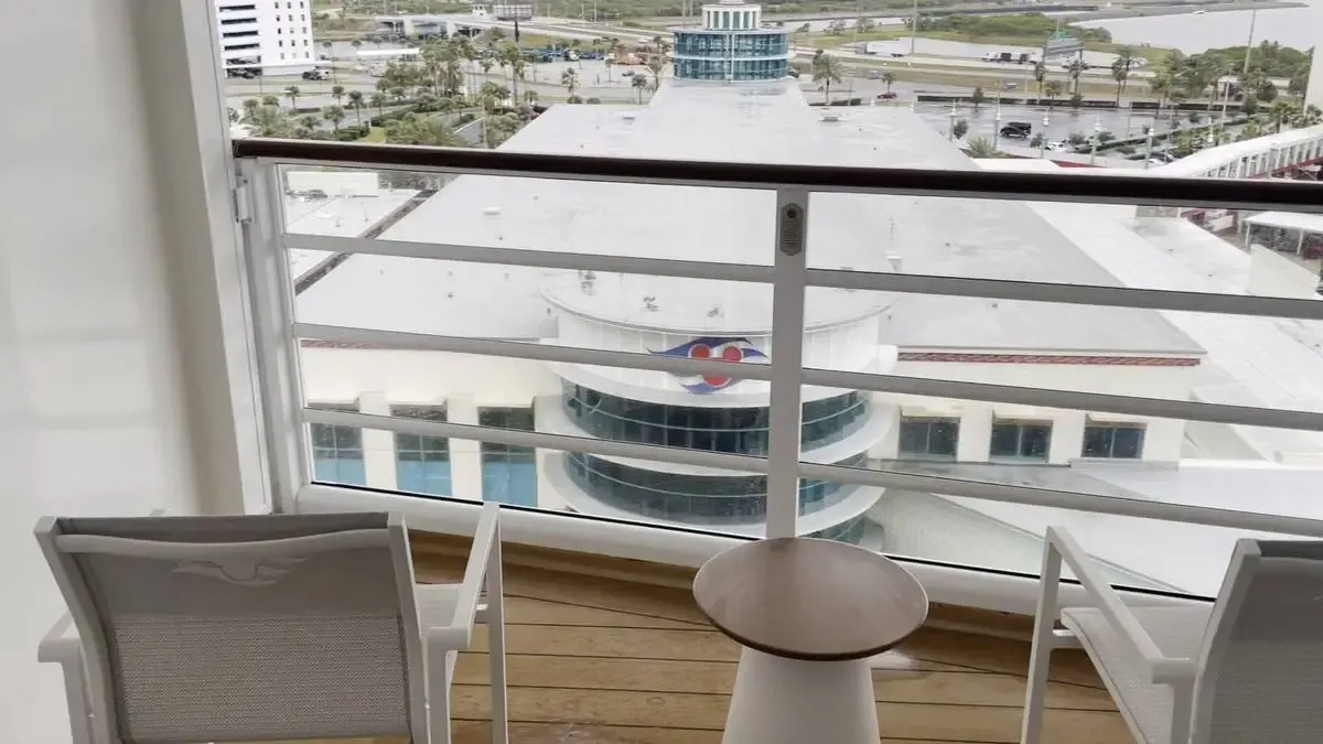Two white chairs and a small round table are on a balcony with a view of a large building with a circular glass section and a logo. The scene includes a distant view of roads and greenery beyond the structure.