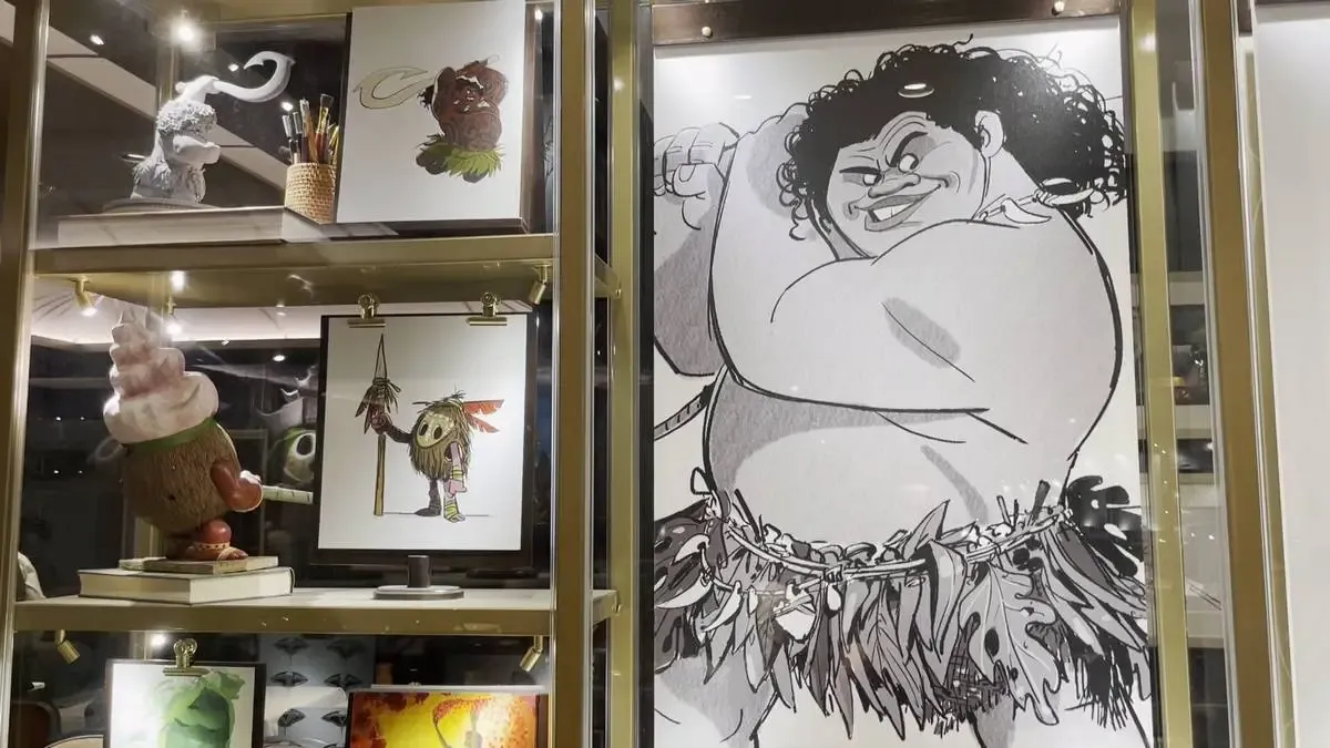 A display case showcases artwork and figurines, including a large black-and-white drawing of Maui from Moana movie. Next to it, shelves hold colorful illustrations and a coconut-themed figurine with a whimsical design. The setting is well-lit, highlighting the vibrant and playful art pieces.