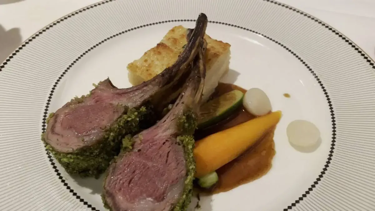 A plate featuring two herb-crusted lamb chops, served with a slice of layered potato gratin, a glazed carrot, and other small vegetables, all arranged elegantly with a rich sauce.