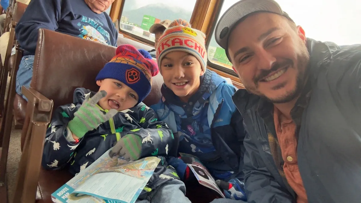 A smiling man takes a selfie with two children on a train. The kids are bundled up in colorful winter hats and jackets, with one holding a map. The train's wooden interior and large windows are visible in the background.