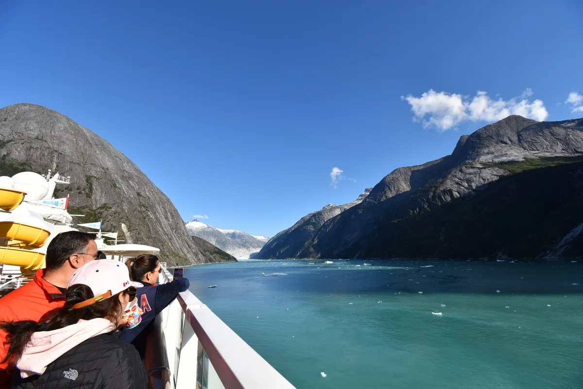 A group of people on a cruise ship deck admire the stunning view of a fjord with clear blue water, surrounded by towering rocky mountains under a bright blue sky.