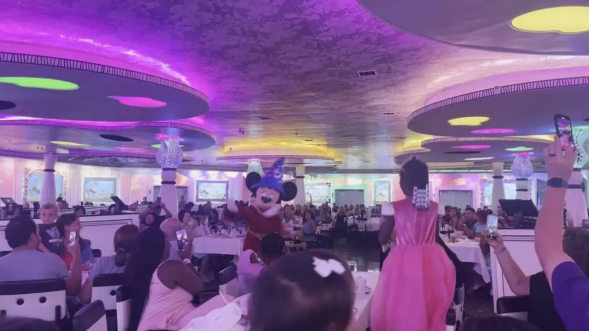 A lively scene in a colorful restaurant, featuring a character in a blue wizard hat entertaining guests. A young girl in a pink dress excitedly approaches, while diners capture the moment on their phones. The ceiling is adorned with vibrant lights, enhancing the festive atmosphere.