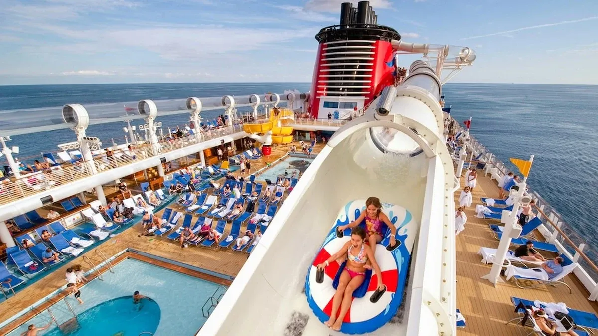 Two children ride a water slide on a cruise ship, surrounded by a vibrant deck with blue lounge chairs and a swimming pool, under a clear blue sky.