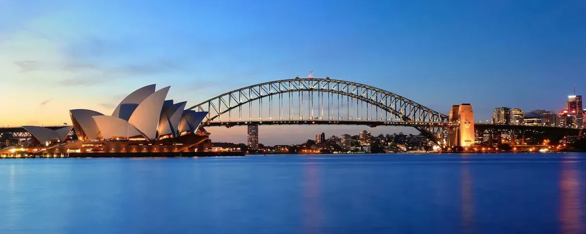 The image shows the iconic Sydney Opera House with its distinctive sail-like structures, set against the backdrop of the Sydney Harbour Bridge at dusk. The city skyline is illuminated, reflecting softly on the tranquil waters of the harbor.