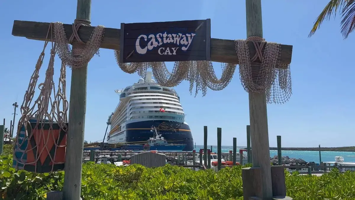 A large cruise ship is docked at Castaway Cay, with a rustic wooden sign and netting in the foreground. The bright blue ocean and clear sky create a tropical setting, and lush greenery surrounds the area.