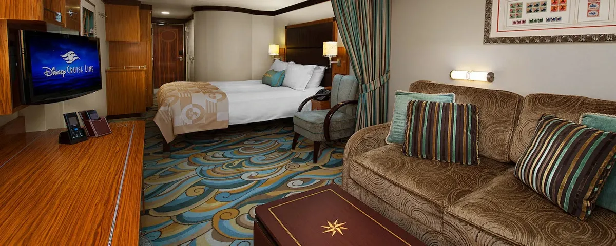 A cozy cruise ship cabin features a comfortable double bed with a patterned beige blanket, a plush brown sofa adorned with striped pillows, and a flat-screen TV displaying "Disney Cruise Line." The room includes a wooden desk, a decorative carpet with swirling patterns, and a framed artwork on the wall, creating a warm and inviting atmosphere.
