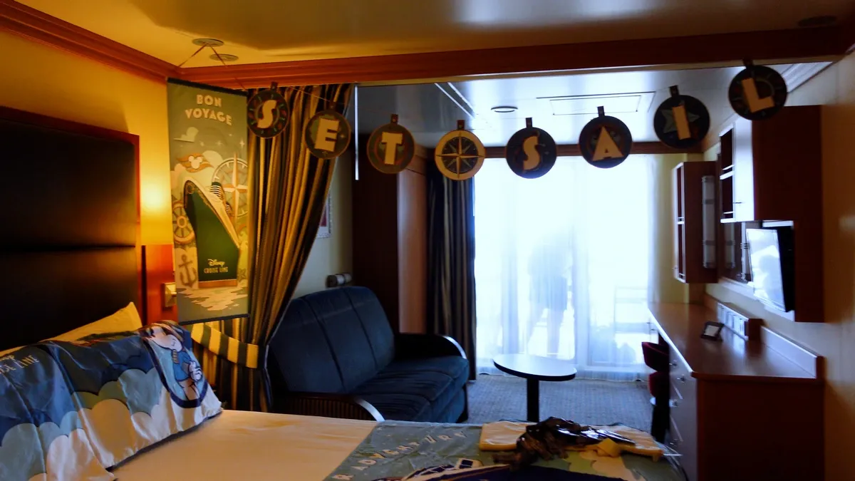 A cozy cruise ship cabin decorated with a "SET SAIL" banner hanging from the ceiling. The room features a bed with themed bedding, a sofa, and a table. A "Bon Voyage" poster is on the wall, and sunlight streams through a glass door leading to a balcony.