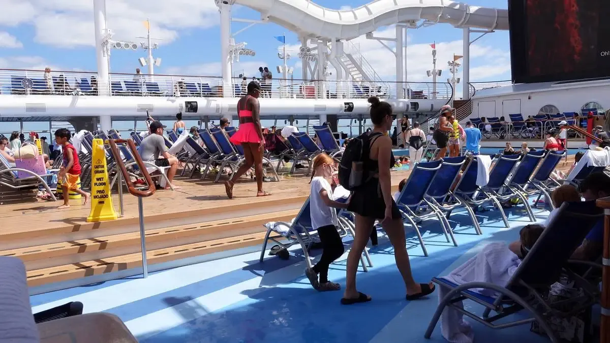 A lively cruise ship deck features people relaxing on blue lounge chairs under a sunny sky. A woman in a bright pink dress walks past, while a child holds hands with an adult nearby. A wet floor caution sign stands on the wooden deck, and a large waterslide and screen are visible in the background.