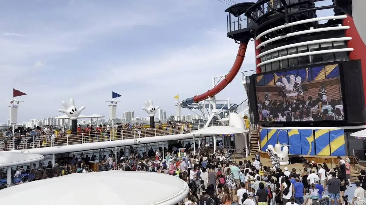 A lively crowd gathers on the deck of a cruise ship, featuring large Mickey Mouse hand decorations and a red water slide. A big screen displays an event, while people enjoy the sunny day with a city skyline in the background.