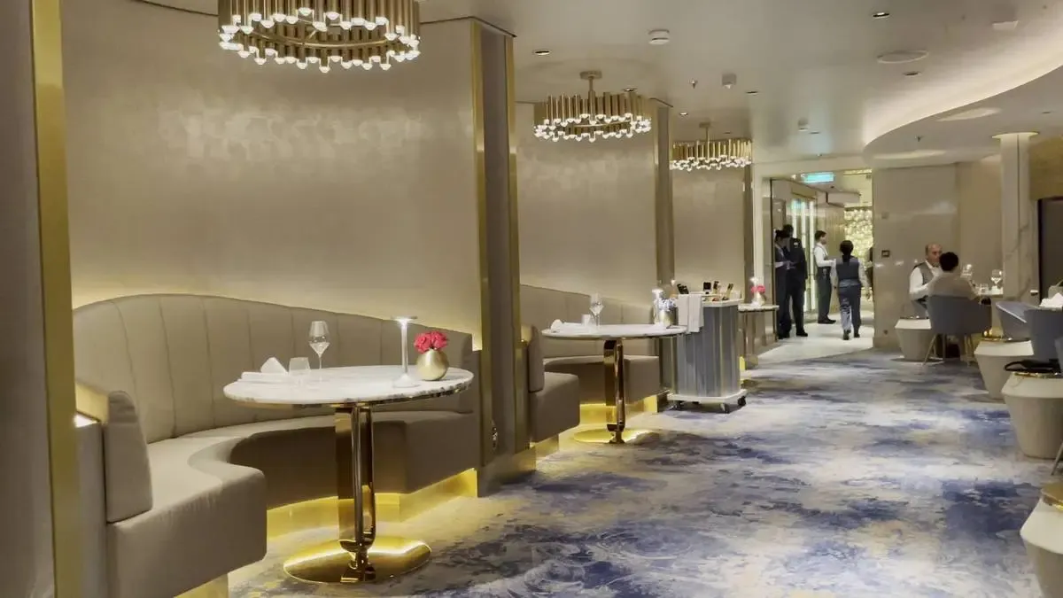 A luxurious restaurant interior featuring elegant curved booths with white marble tables and golden accents. The floor is covered in a stylish blue and beige patterned carpet, and modern chandeliers hang from the ceiling, casting a warm glow. A few patrons and staff are visible in the background, enhancing the upscale ambiance.