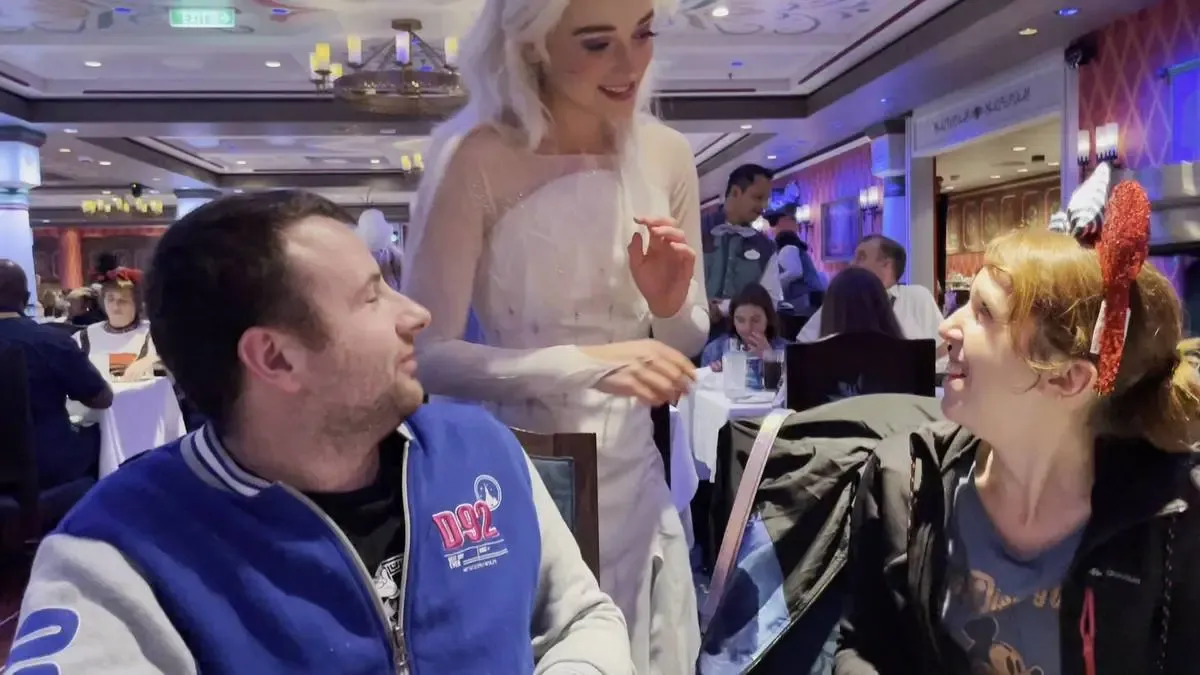 A woman dressed as princess Elsa interacts with two seated adults in a lively restaurant setting. The man wears a blue varsity jacket, while the woman sports red glittery Minnie Mouse ears. The background features other diners and warm lighting.