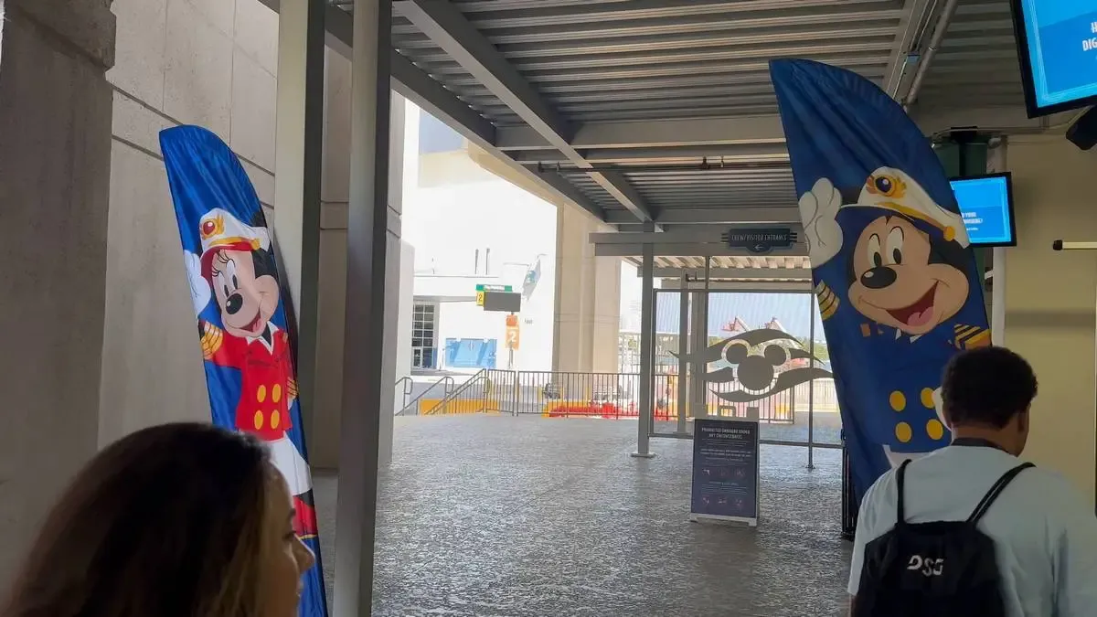Two large banners featuring Mickey and Minnie Mouse in a sailor outfit are displayed in an open-air terminal area. People are walking nearby, and a gate with a similar cartoon design is visible in the background.