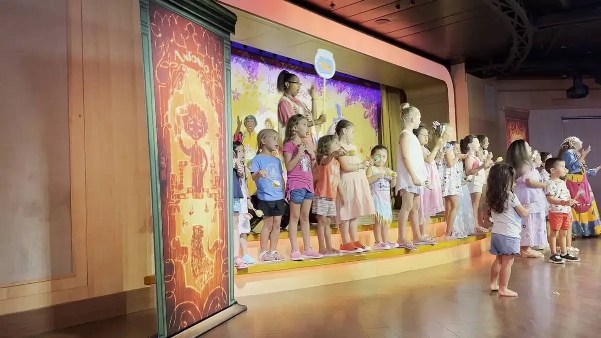 A group of children stand on a stage, participating in a colorful performance. They are dressed in casual summer clothes, with a backdrop featuring vibrant artwork. The setting appears lively and engaging, with a mix of boys and girls attentively involved in the activity.