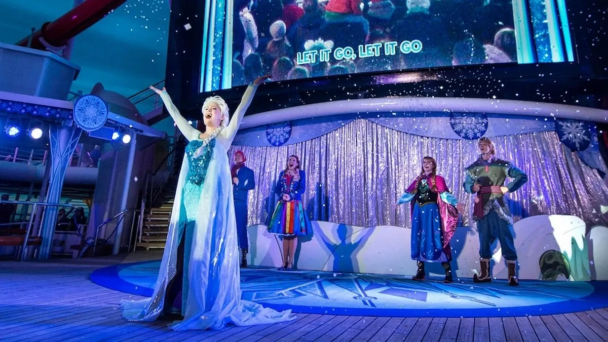 Performers dressed as characters from Frozen sing on a stage decorated with snowflakes, under a large screen displaying "Let it Go." The scene is lively, with artificial snow falling and colorful lighting enhancing the winter-themed atmosphere.