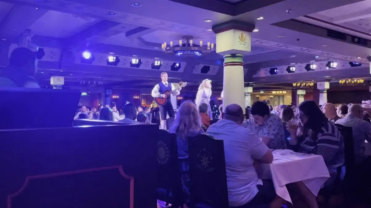 A musician plays guitar on a stage in a dimly lit restaurant, surrounded by dining guests seated at tables with white tablecloths. The room features decorative columns and ceiling lights, creating a lively atmosphere.