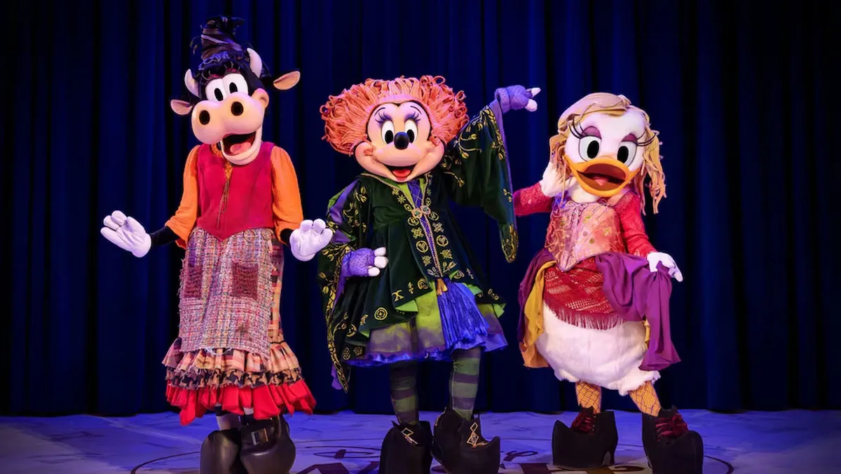 Three costumed characters stand on stage in front of a dark blue curtain, each wearing vibrant Halloween-themed outfits; the left character is dressed in a colorful patchwork skirt, the middle one in a green and purple witch outfit, and the right one in a pink and red ensemble.