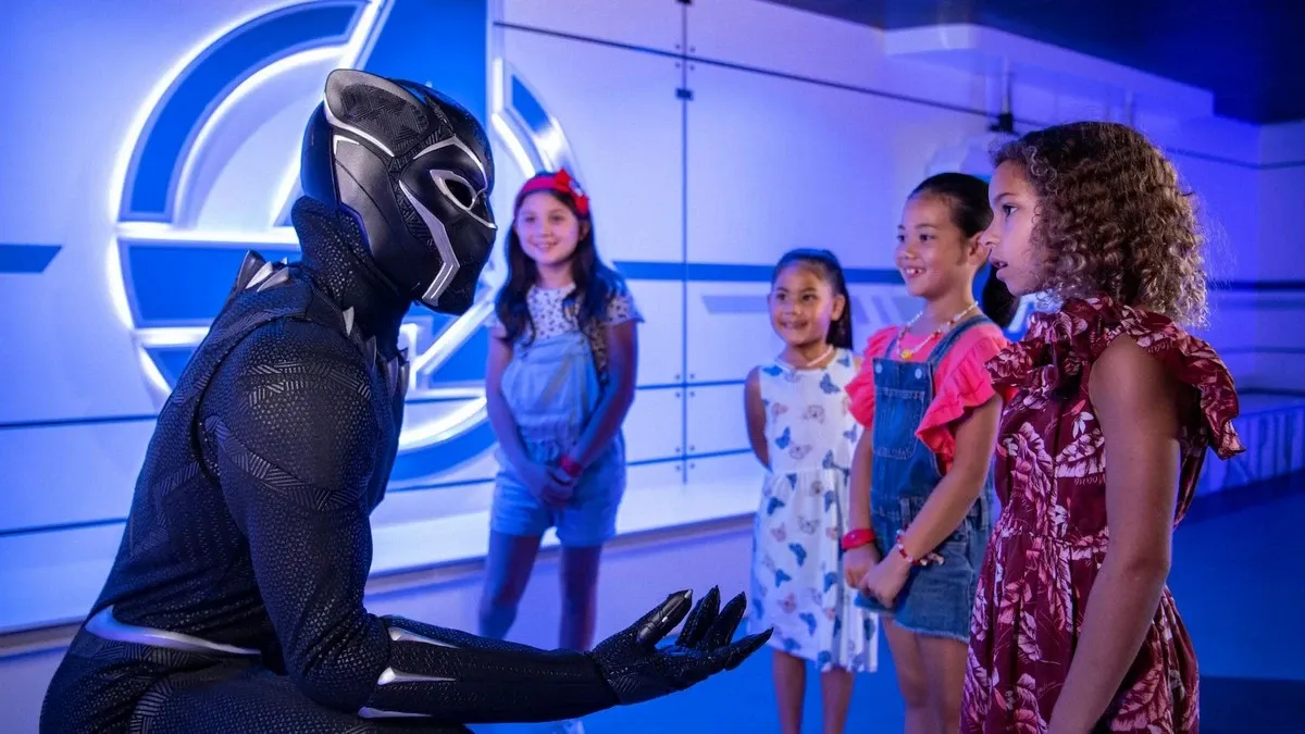 A person in a Black Panther costume interacts with four smiling children in a brightly lit room with a futuristic design. The children, dressed in colorful outfits, appear engaged and excited, standing near a large illuminated symbol on the wall.