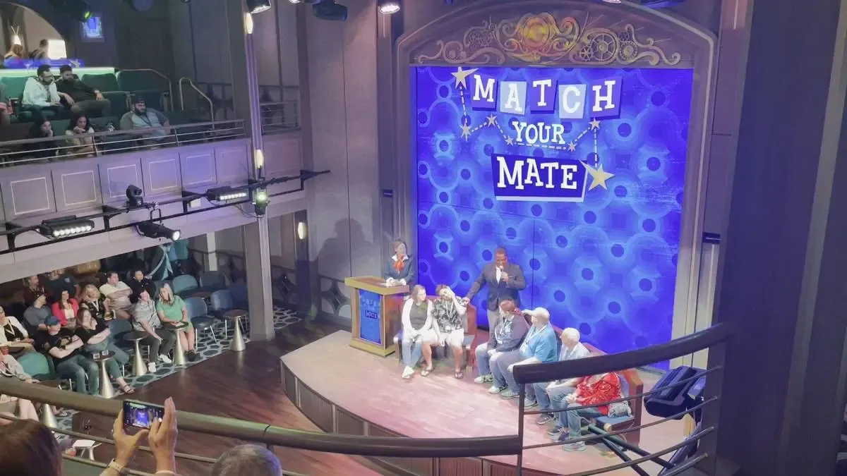 A lively game show titled "Match Your Mate" is taking place on a stage with a bright blue backdrop. Several contestants are seated on stage, while a host stands nearby with a microphone. The audience is watching from tiered seating, and a person in the foreground is capturing the moment with a phone camera.