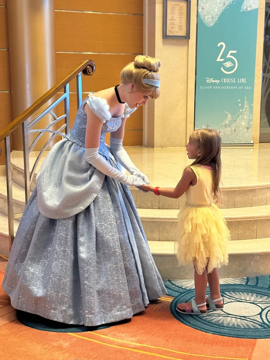 A woman dressed as a princess in a sparkling blue gown and gloves bends down to greet a young girl in a yellow dress. They are on a cruise ship, with a "Disney Cruise Line Silver Anniversary at Sea" sign in the background.