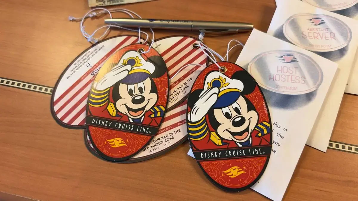 Disney Cruise Line luggage tags featuring a cheerful cartoon character in a captain's uniform are displayed on a wooden surface. A pen and some paperwork, including a "Host Hostess" card, are visible in the background.