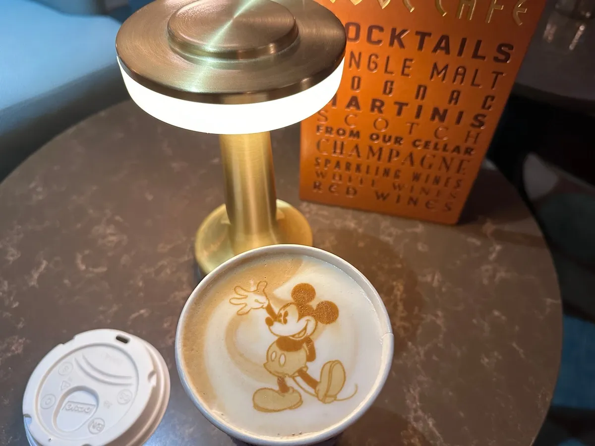 A cup of coffee on a marble table features latte art of a cartoon mouse character. Nearby, there's a modern brass table lamp and a menu with drink options like cocktails and champagne.