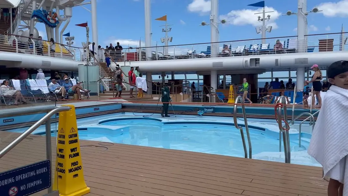 A cruise ship deck features a small pool surrounded by people lounging and walking, with some wrapped in towels. Bright yellow caution signs indicate a wet floor. The deck is bustling with activity under a clear blue sky, with additional seating and people visible on the upper level.