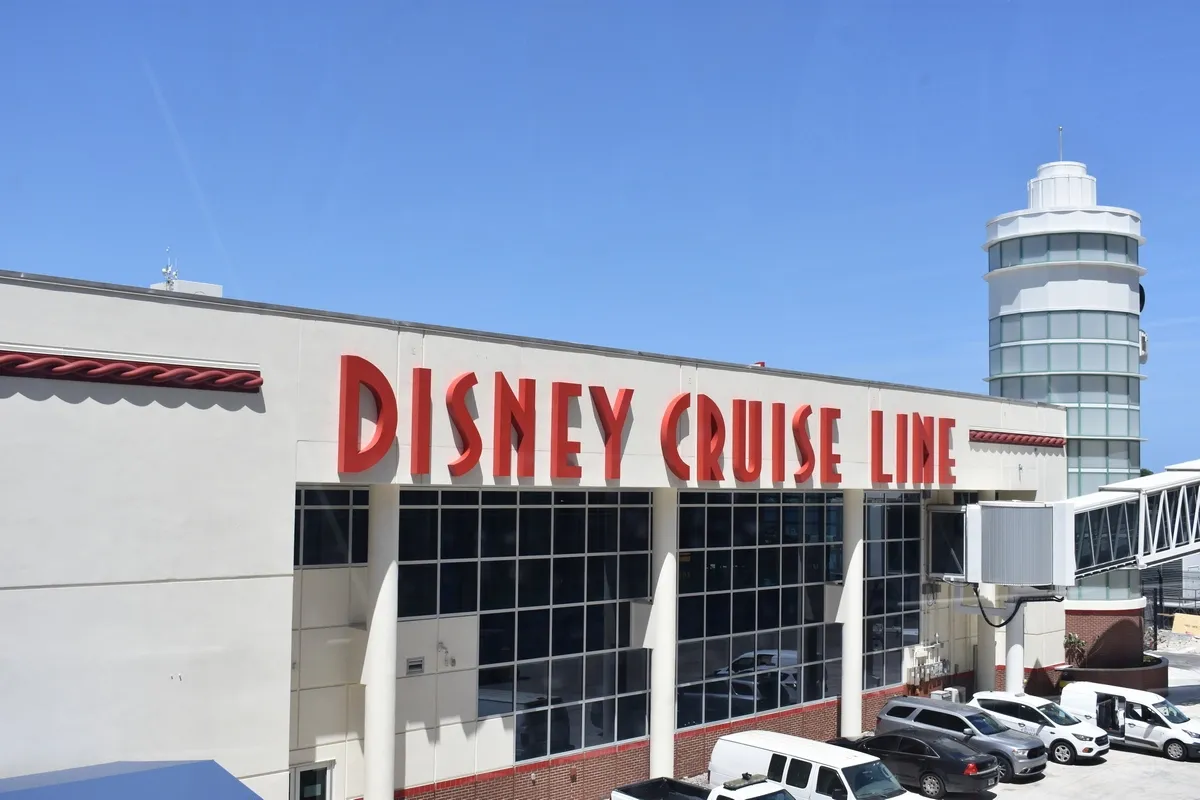 The image shows a Disney Cruise Line terminal with large red lettering on the building. The structure features a modern design with large windows and a cylindrical tower in the background. Several white vehicles are parked in front of the terminal under a clear blue sky.