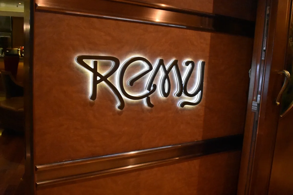 Illuminated sign reading "Remy" in elegant script on a warm brown wall, with a polished wooden frame and a glimpse of a cozy interior setting.