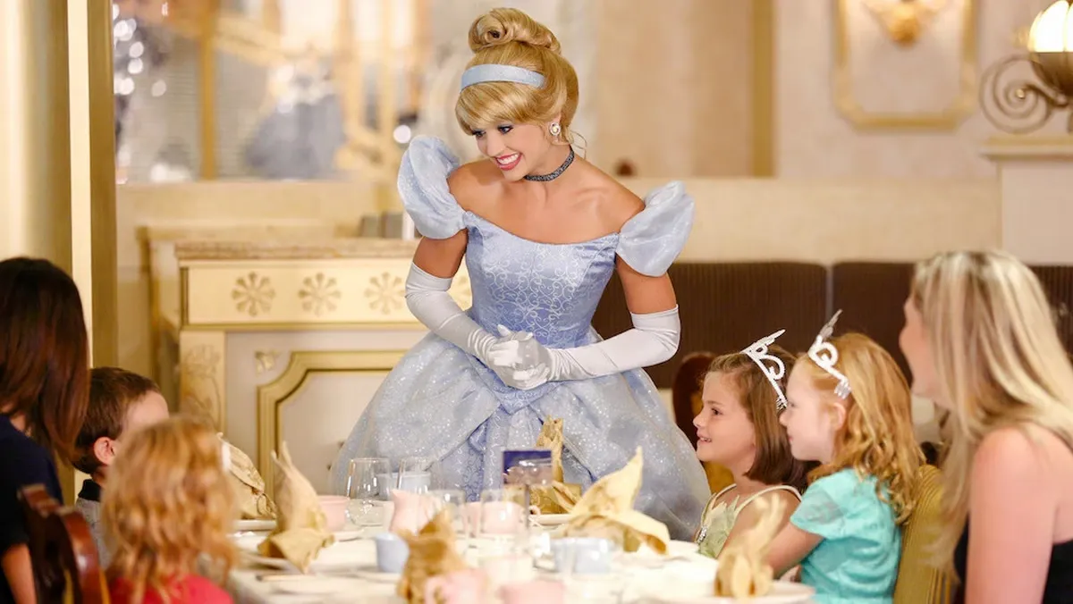 A person dressed as Cinderella, wearing a sparkling blue gown and white gloves, smiles warmly at a group of children seated around a dining table. The children, some wearing tiaras, appear delighted in the elegant, ornate setting.