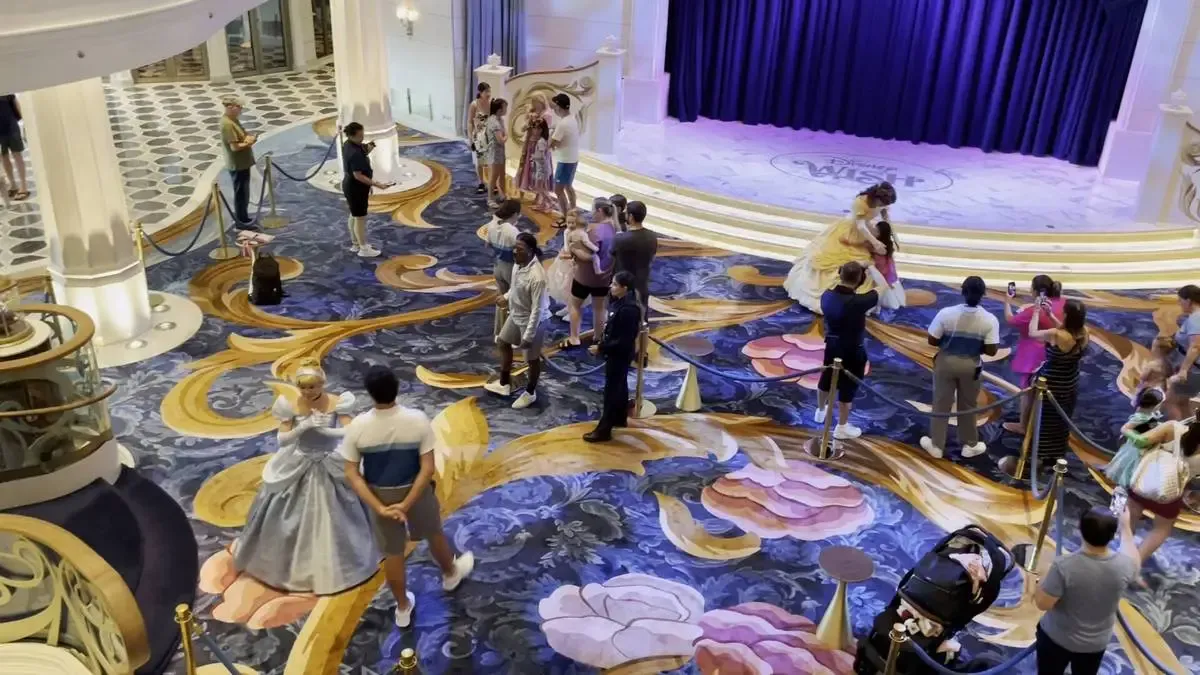 People are gathered in an ornate lobby with a blue and gold patterned carpet, meeting characters in elaborate costumes. A woman in a yellow gown hugs a child near a small stage, while another in a blue dress interacts with a visitor. The scene is lively, with visitors taking photos and children dressed in costumes.