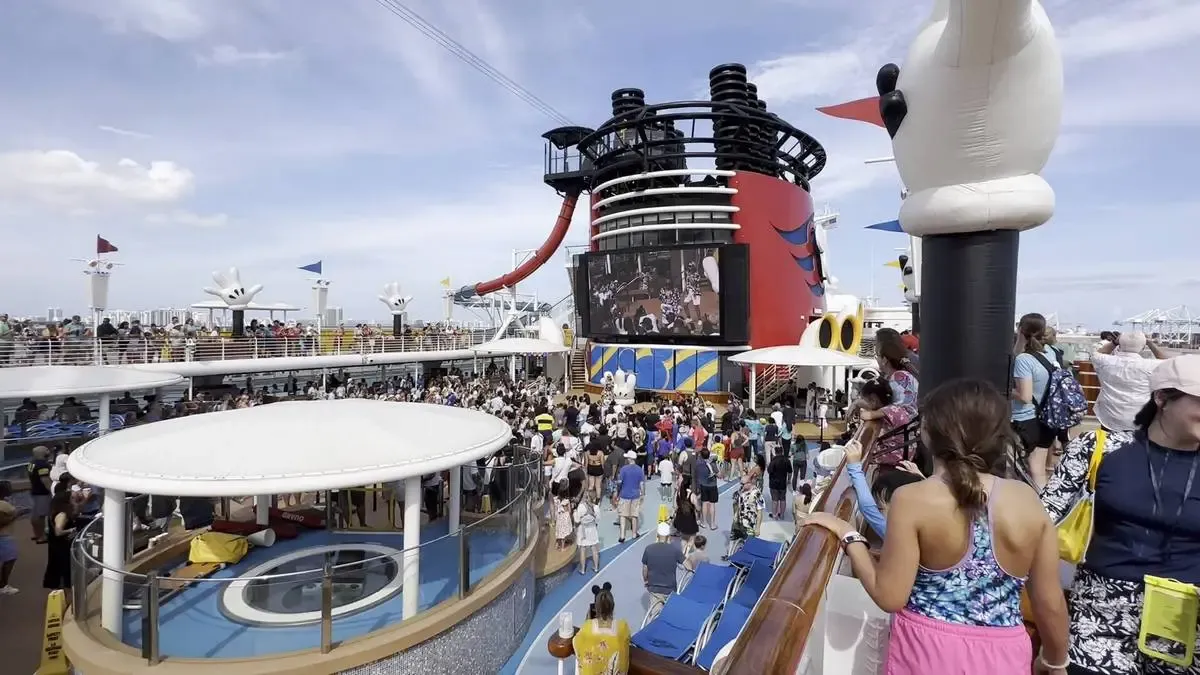 A lively crowd gathers on the deck of a cruise ship, featuring a large screen and colorful water slide. People are enjoying the sunny weather, surrounded by various deck amenities and decorations, including inflatable figures. The atmosphere is vibrant and festive.