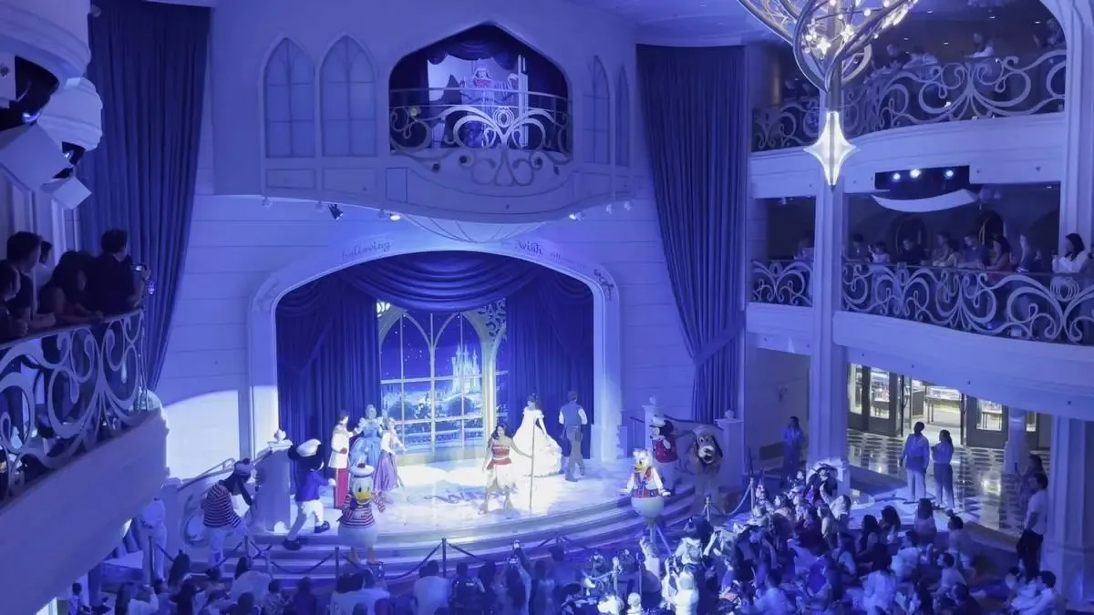 A vibrant stage performance features costumed characters in front of an audience, set in a grand, elegantly decorated theater with balconies. The scene is bathed in blue lighting, highlighting the ornate architecture and a large window backdrop depicting a castle.