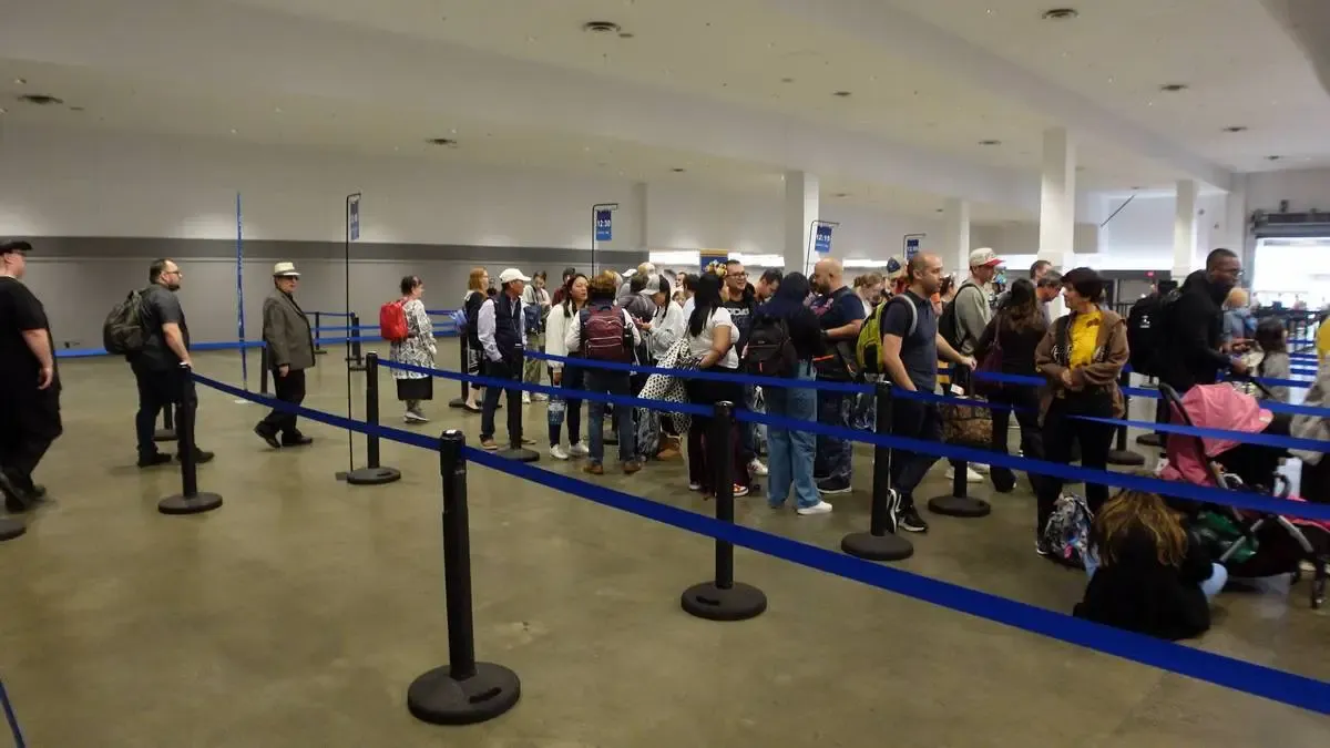A group of people stands in line at an indoor venue, separated by blue stanchions. The crowd includes individuals with backpacks and strollers, suggesting a travel or event setting. The space is spacious with a concrete floor and overhead signs.