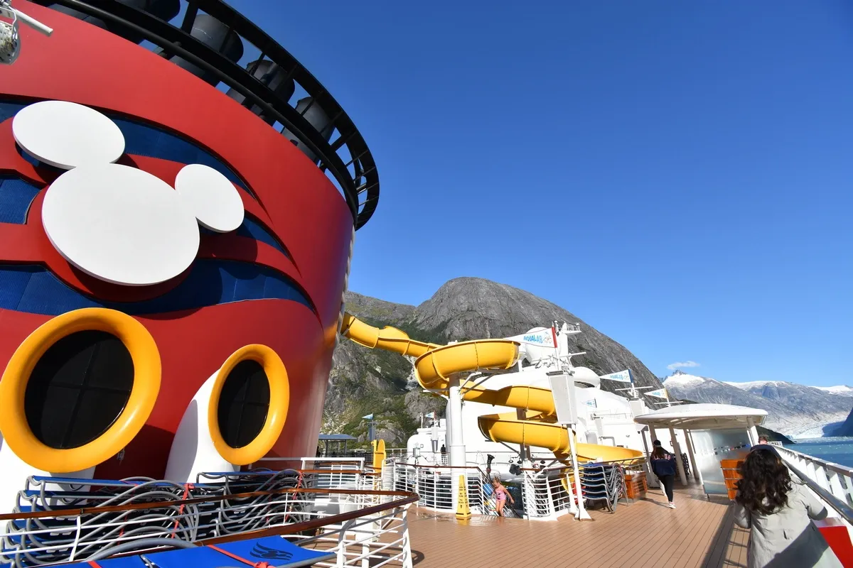 A cruise ship deck features a large red and blue funnel with a Mickey Mouse logo, alongside a vibrant yellow waterslide. The ship is set against a backdrop of majestic mountains and a clear blue sky, with a few people enjoying the sunny day on deck.
