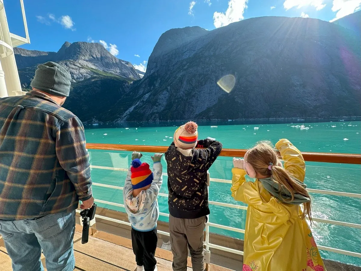 A man and three children, all dressed warmly, stand on a ship's deck gazing at the stunning turquoise waters and towering mountains in the background. The children wear colorful hats and look over the railing, captivated by the scenic view under a bright, sunny sky.