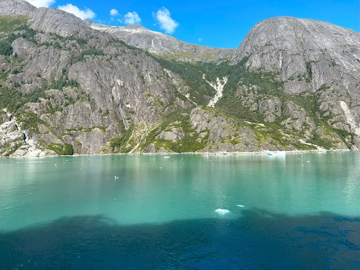A serene turquoise lake is surrounded by rugged, rocky mountains with patches of greenery under a clear blue sky.
