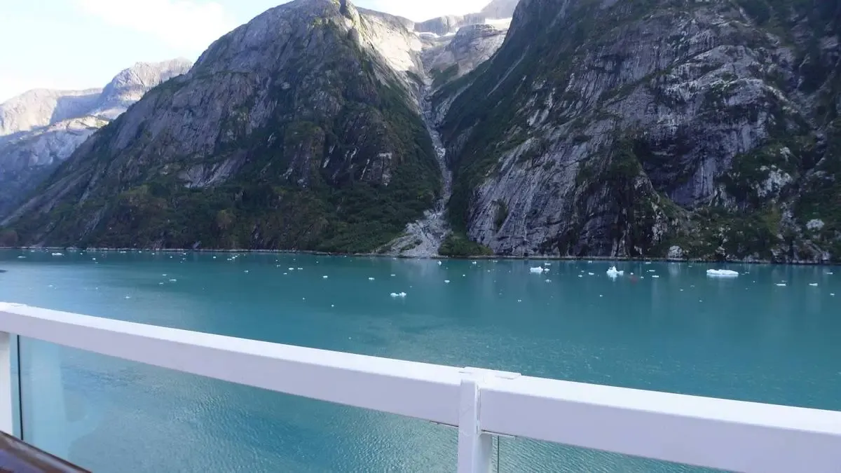 A scenic view of a turquoise fjord surrounded by towering, rugged mountains with patches of greenery. Small icebergs float on the water, and a white railing is visible in the foreground, suggesting the photo was taken from a boat or a balcony.