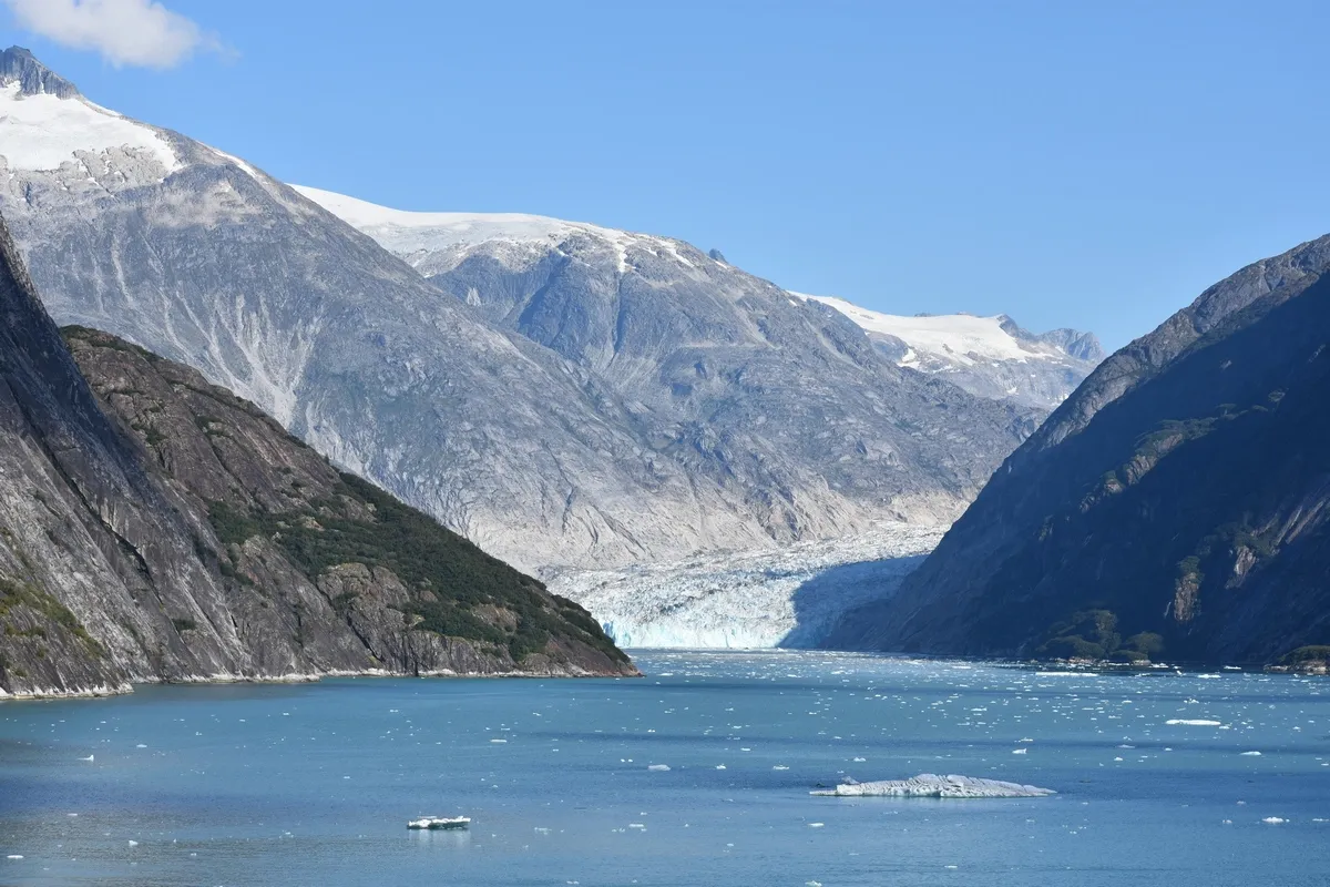 A breathtaking view of a glacier nestled between rugged, snow-capped mountains, with a serene blue fjord in the foreground dotted with small icebergs under a clear blue sky.