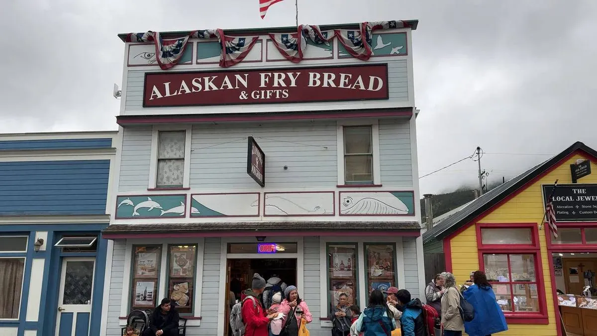 A group of people stand outside a quaint, two-story building with a sign reading "Alaskan Fry Bread & Gifts." The building is adorned with decorative bunting and features a light blue facade with white and red accents. Nearby, colorful neighboring shops add to the charming street scene under a cloudy sky.