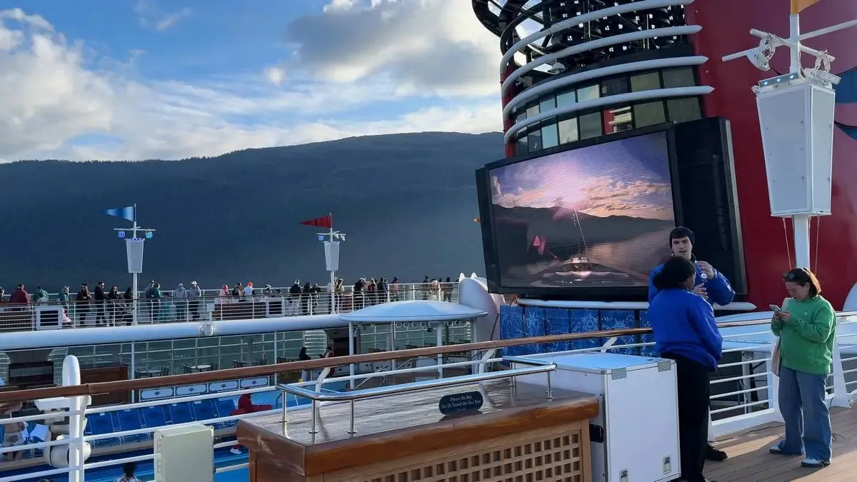 People are gathered on the deck of a cruise ship featuring a large screen displaying a scenic image of a sunset over water, with a mountainous landscape in the background under a partly cloudy sky.