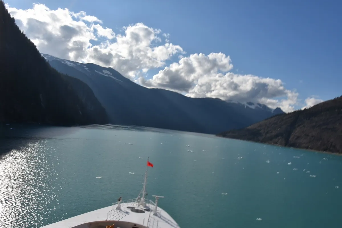 A ship's bow glides through a serene, turquoise fjord surrounded by towering, shadowy mountains under a bright blue sky filled with scattered clouds.