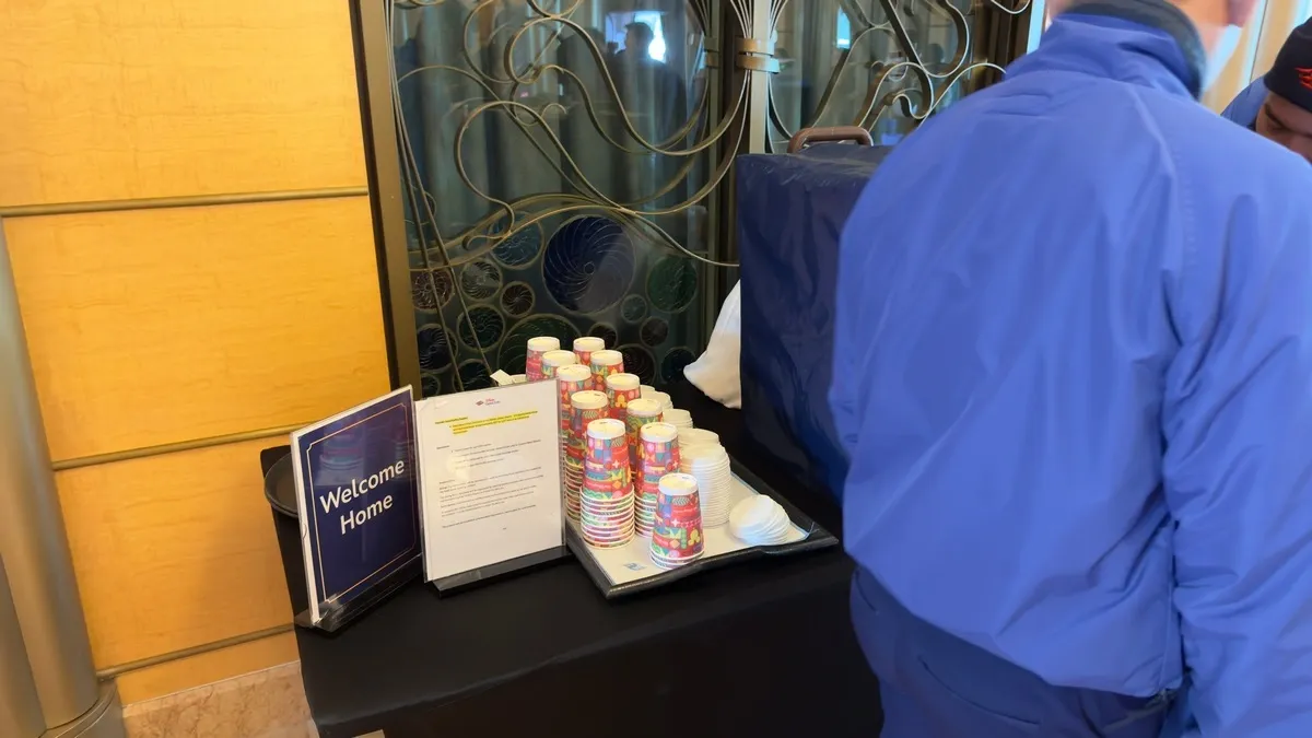 A person in a blue jacket stands next to a table with a "Welcome Home" sign, a stack of colorful paper cups, and a covered container, likely for refreshments. The setting appears to be indoors, with decorative glass in the background.