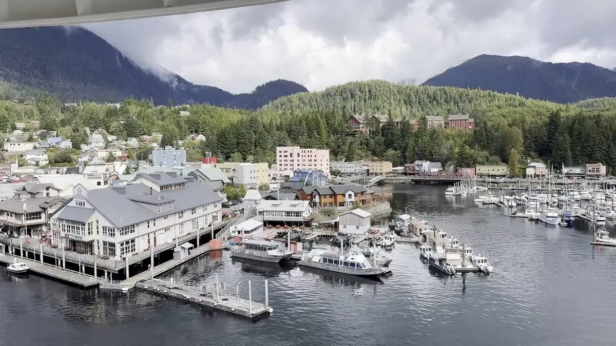 A scenic harbor view features several boats docked along piers, with charming buildings lining the waterfront. In the background, lush green forests cover rolling hills beneath a cloudy sky, creating a picturesque backdrop.