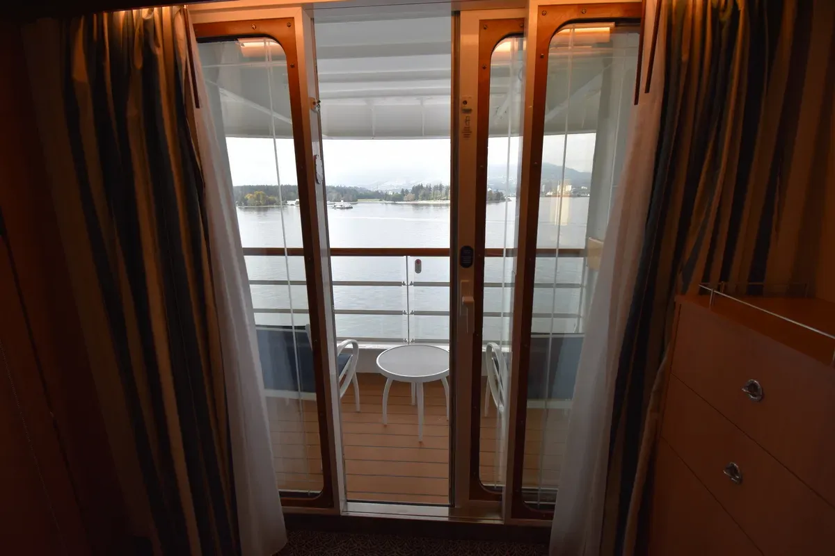 A view from a room showing sliding glass doors leading to a balcony with a small round table and chairs. Beyond the balcony, there's a scenic view of a calm body of water with distant land and boats. The room features striped curtains and a wooden dresser.