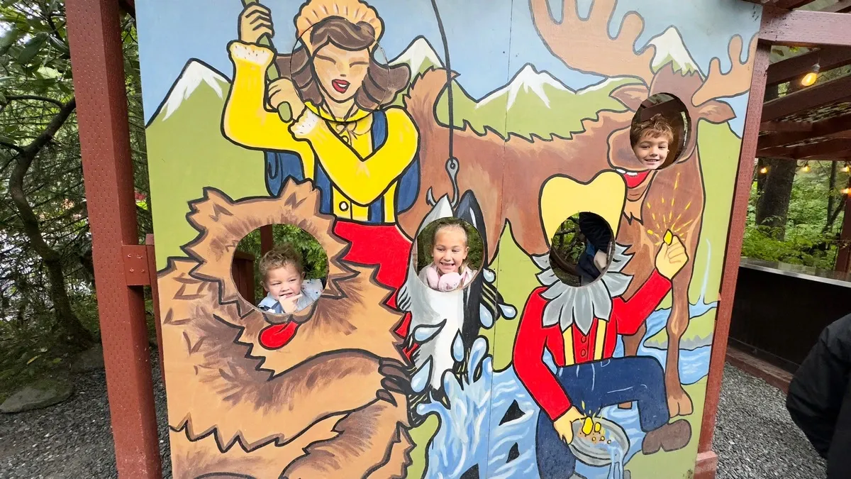 Three children pose with their faces through cutouts in a colorful outdoor mural depicting a lively wilderness scene, featuring a fisherwoman, a bear, and a prospector.