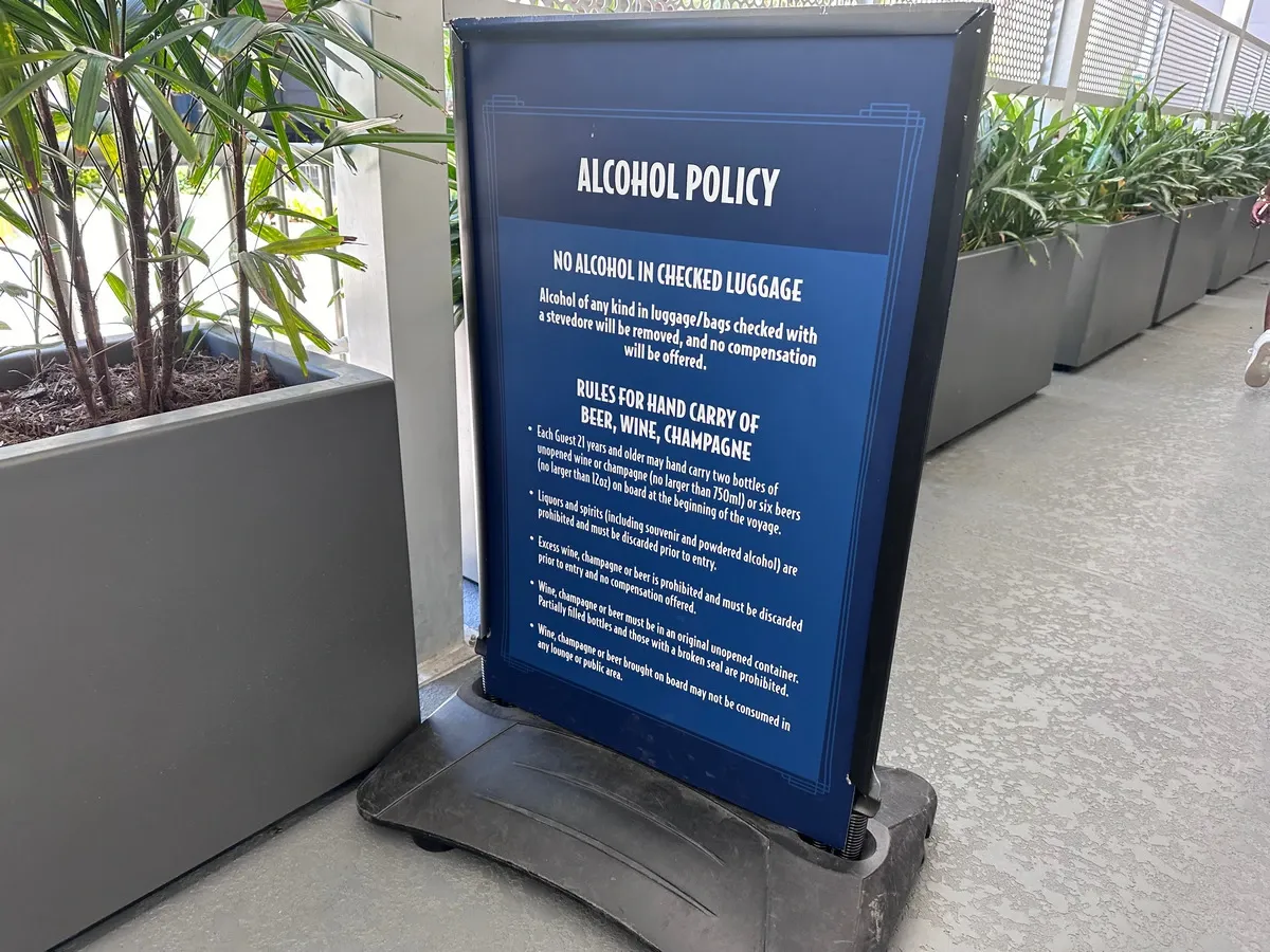 A sign detailing the alcohol policy stands on a concrete floor beside large potted plants. The sign outlines rules against alcohol in checked luggage and specifies limits for hand-carrying beer, wine, and champagne. The text is white on a blue background.