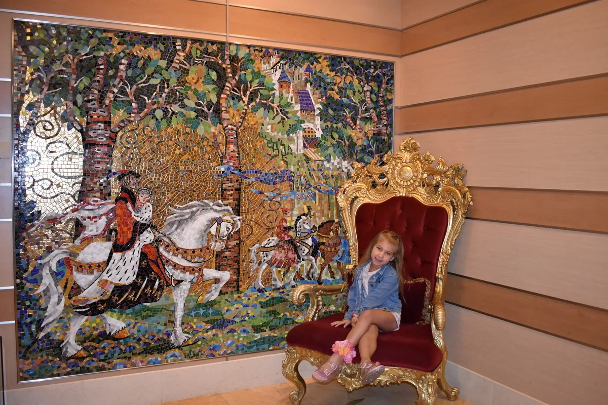A young girl sits happily on an ornate, gold and red throne in front of a vibrant mosaic mural depicting a medieval scene with knights on horseback amidst a forest.